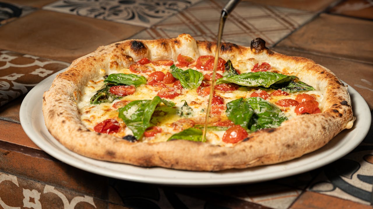 Delicious Neapolitan pizza with cheese, tomatoes, and fresh basil being drizzled with olive oil.